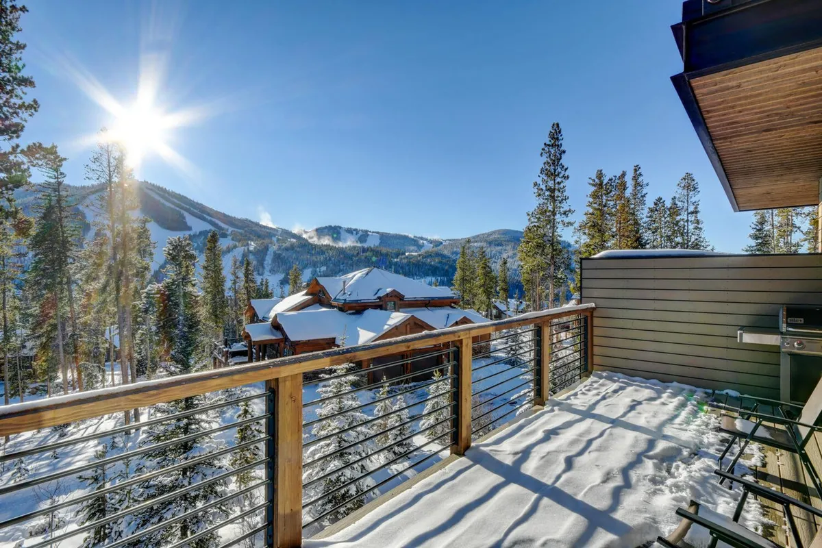 A snow-covered balcony framed by pine trees and distant mountains, sunlight streaming through the trees. The space offers direct outdoor access from the private deck, perfect for quiet mornings or stargazing at night.