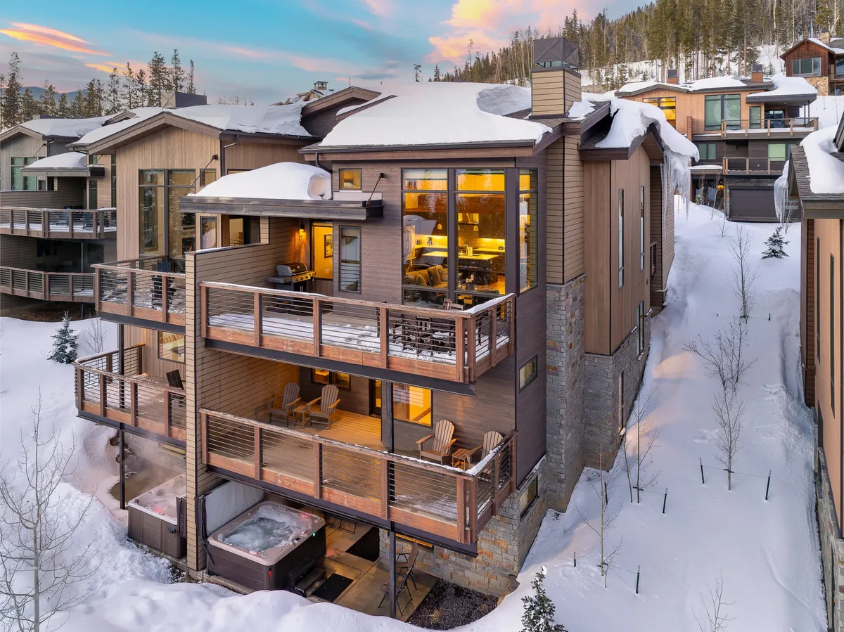 The image showcases the exterior of a luxurious single-family villa set against the snow-covered landscape of Winter Park, Colorado. A private hot tub steams invitingly on the deck, surrounded by comfortable seating and a spacious balcony perfect for gatherings. Expansive windows frame panoramic mountain views, while hints of sunset color the sky, promising serene evenings and memorable moments.