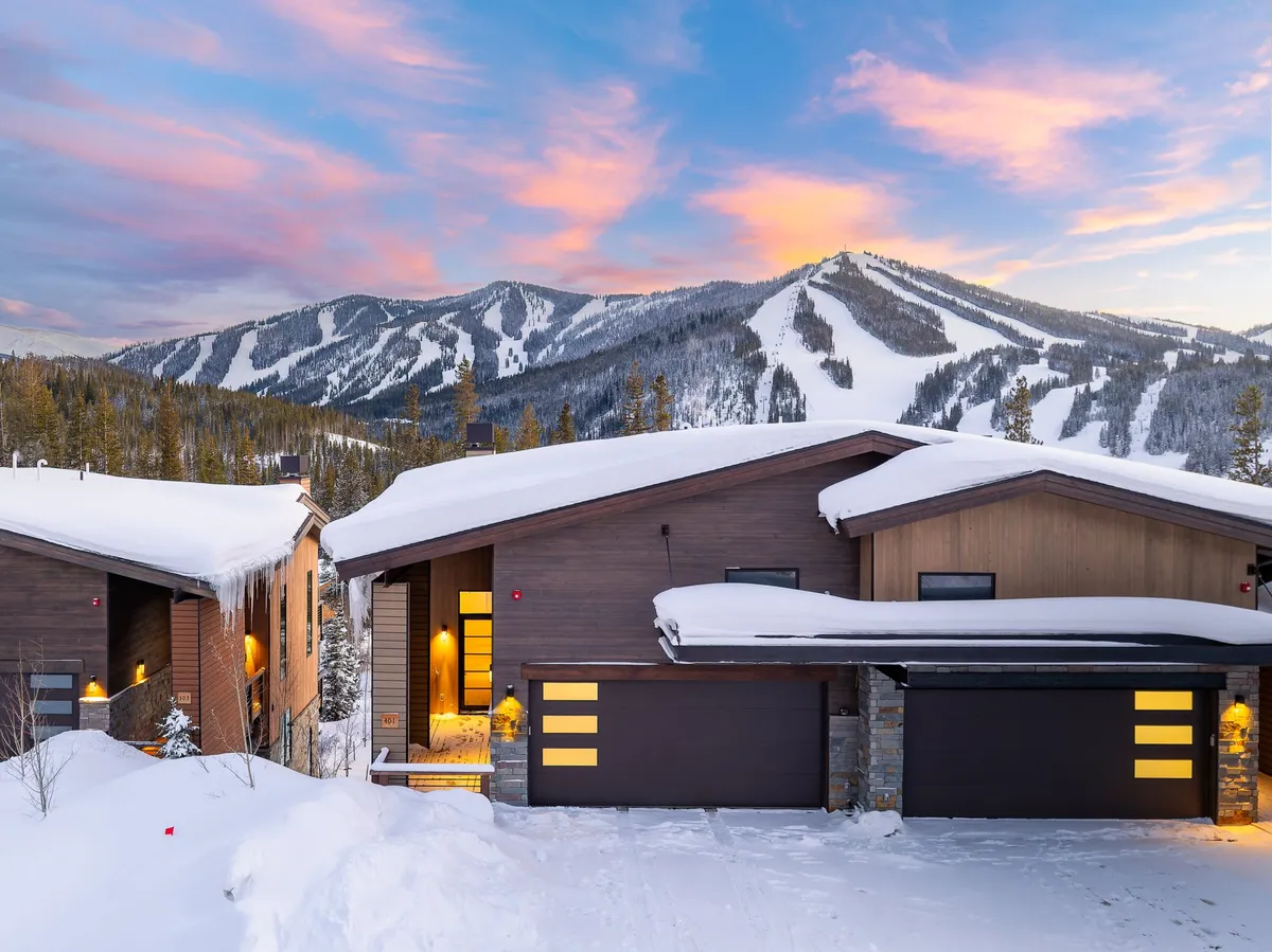 An exterior shot of the home reveals modern architecture nestled in a snow-covered landscape with mountain views, highlighted by a warmly lit entrance. The scene exudes a welcoming, cozy vibe that hints at the adventures and relaxation that await inside, while also placing the property in its scenic context.