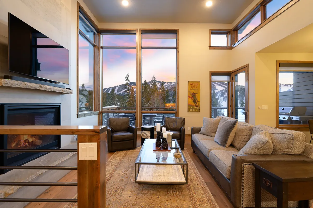 This image showcases a spacious living area with large windows framing sweeping mountain views. There is a cozy seating arrangement near a stone fireplace, with a coffee table set in the center. The natural wood beams overhead and the decor reinforce a refined Colorado mountain style. Ample daylight pours into the room, highlighting the inviting and serene atmosphere.