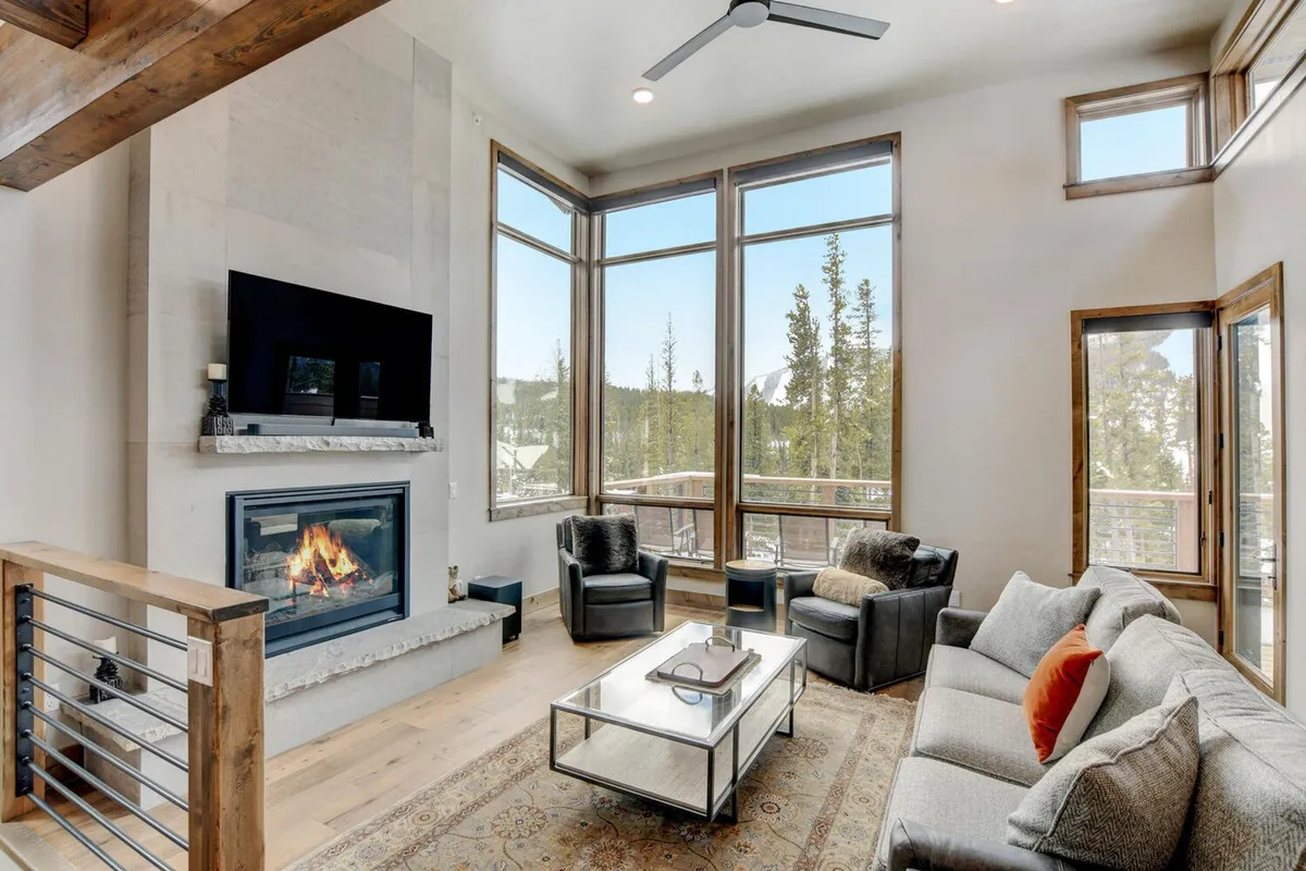 A second angle of the main living area highlights a large seating area gathered around a modern fireplace, with expansive windows letting in sunlight and offering exterior views. The stylish coffee table and decorative rug add warmth, while the setup is ideal for socializing and family time.