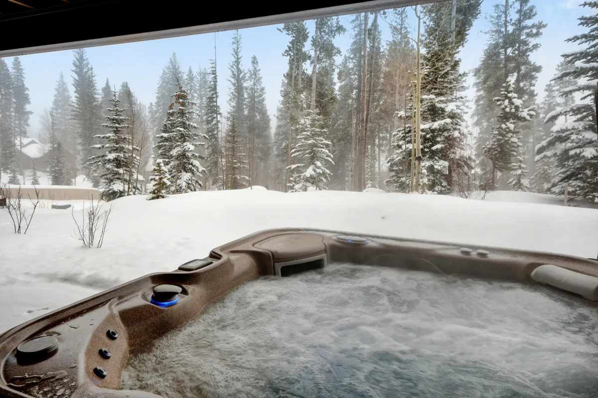 An inviting outdoor hot tub nestled amidst snow-covered trees, with a distant cabin visible. The wintry landscape and the steam rising from the tub evoke relaxation and luxury—a quintessential après-adventure retreat.