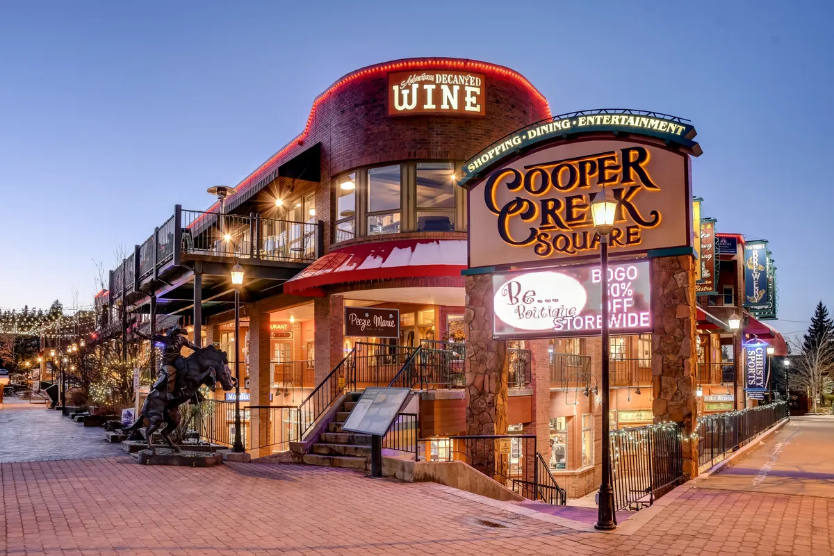 This exterior image features the festive Cooper Creek Square, with prominent signage, a wine shop, outdoor seating areas, decorative lighting, and a distinctive horse sculpture. The scene suggests opportunities for local exploration, group dining, and social outings.