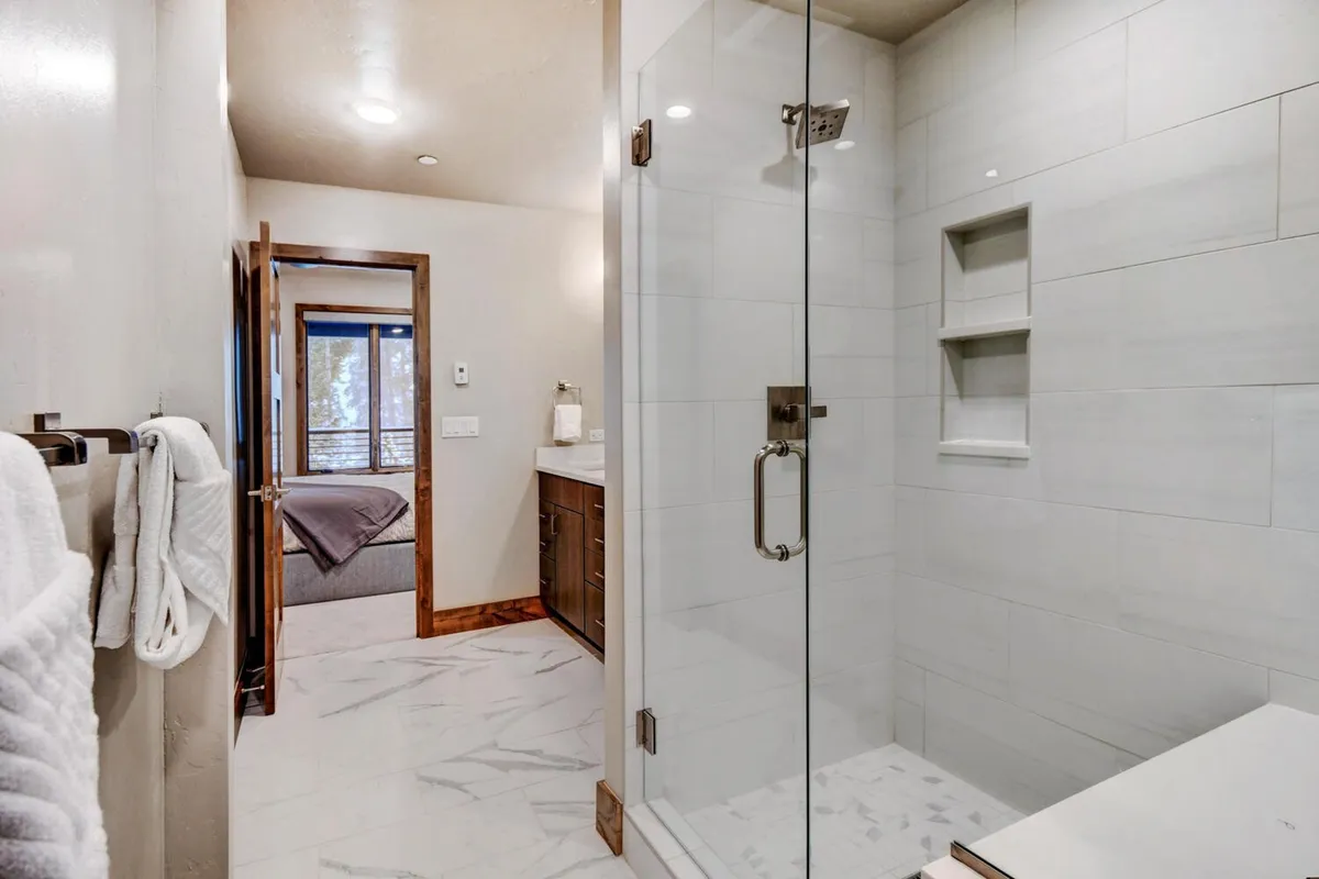 A calming en suite bathroom with a glass shower enclosure, dark wood vanity, and a large window offering views and natural light. The space feels spa-like and luxurious, echoing the master suite description.