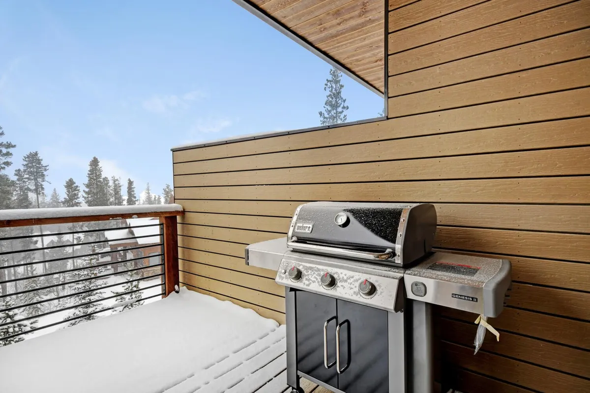 An exterior deck space featuring a modern grill set against a wooden railing, overlooking a snow-covered balcony. The setting is inviting for year-round barbecues, with space to relax and enjoy the winter landscape while preparing outdoor meals.