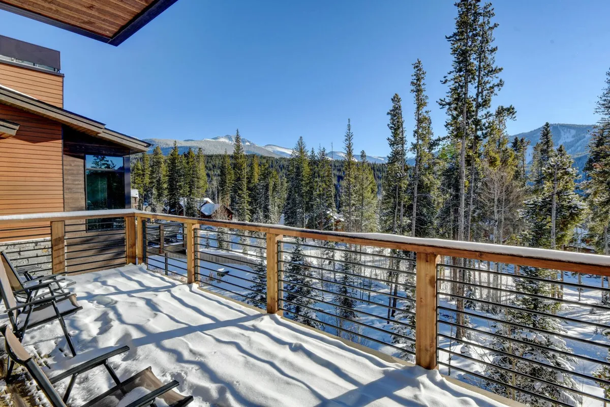 A snow-covered balcony with a wooden railing overlooks surrounding pine trees and a breathtaking view of the mountains. The tranquil setting invites early morning coffee, reading, or wildlife watching, with the promise of stunning sunrises over the Rockies.