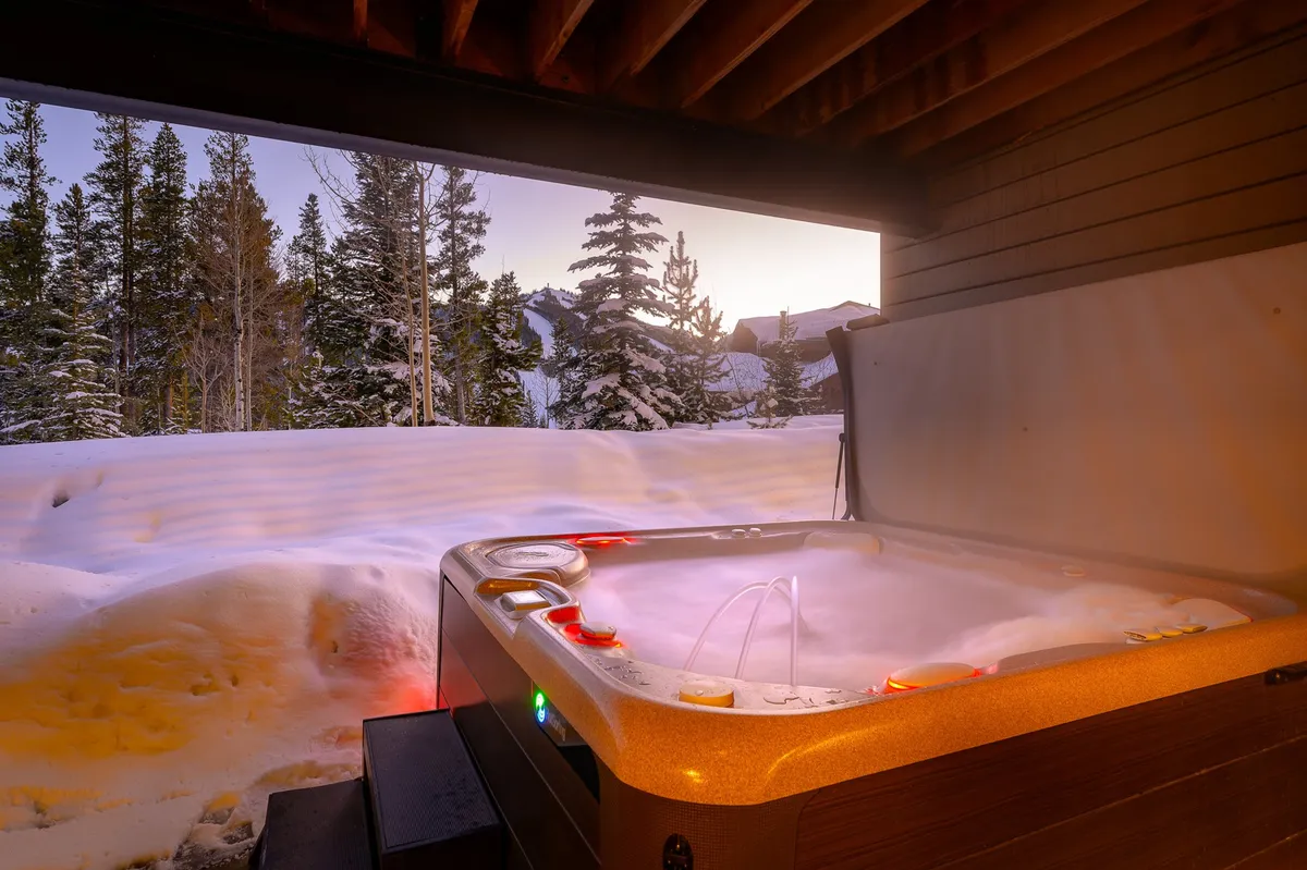 An inviting exterior hot tub set against a snow-covered landscape, surrounded by trees and nature. The serene scene captures the essence of relaxation after a day outdoors, with the hot tub steaming in the cold mountain air.